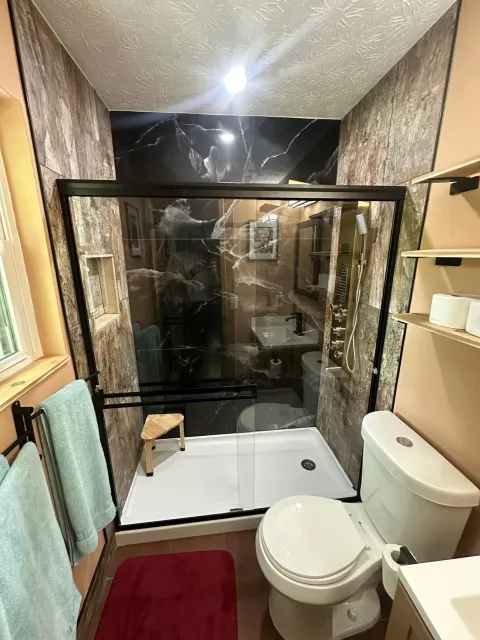 Compact bathroom with glass shower, toilet, sink, and red bath mat.