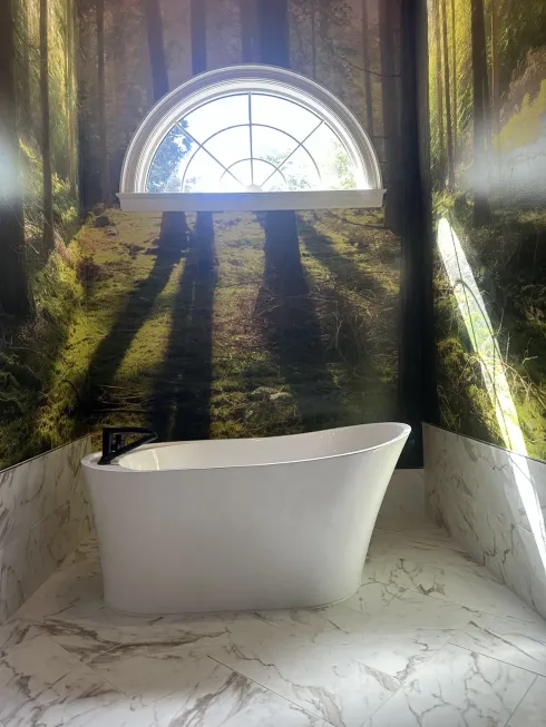 Small bathtub under a sunlit arched window with forest mural walls and marble-patterned floor