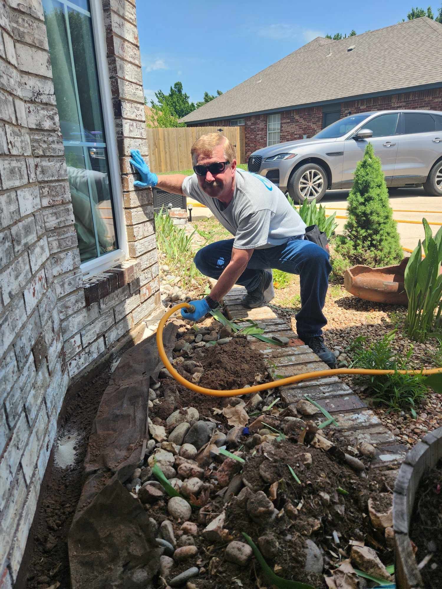 A man is kneeling down in front of a brick building with a hose.
