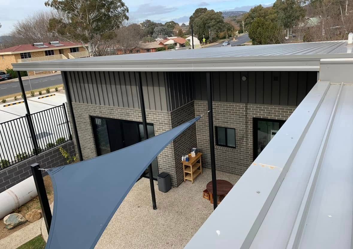 Courtyard of a modern building with shade sail, gravel, and dark gray exterior siding. — Your Craftsmen in Eleebana, NSW