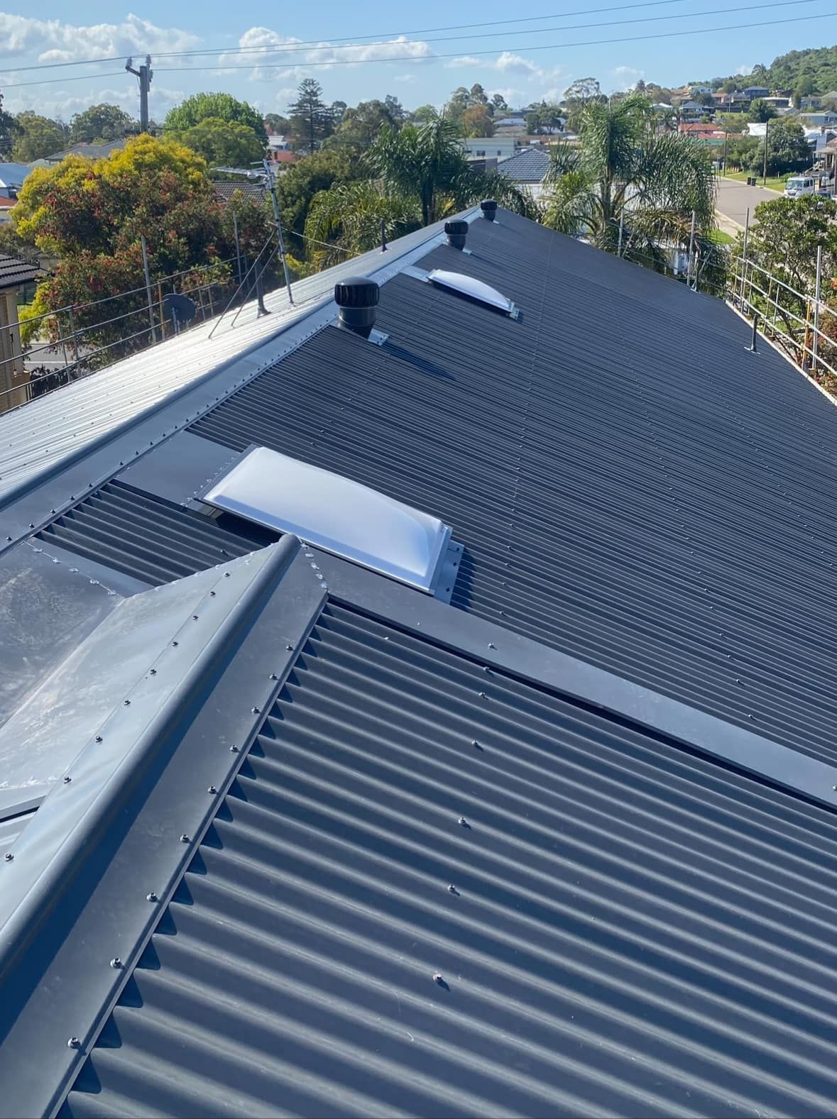 Dark Gray Corrugated Metal Roof With Skylights and Vents, Set Against a Cityscape Backdrop — Your Craftsmen in Eleebana, NSW