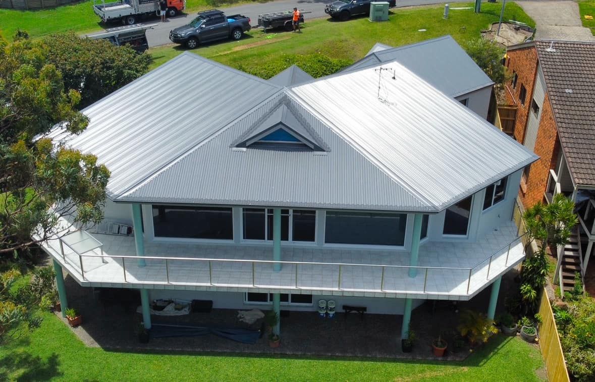 House with a white exterior, silver roof, and wraparound deck. Overlooks green grass, cars, and a building on the right. — Your Craftsmen in Eleebana, NSW