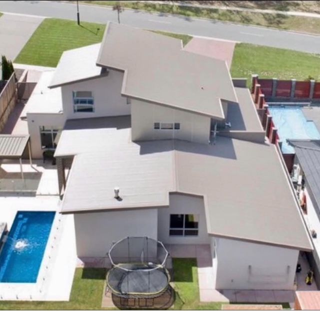 Aerial view of a two-story beige house with a tan metal roof, featuring a backyard pool and a trampoline.