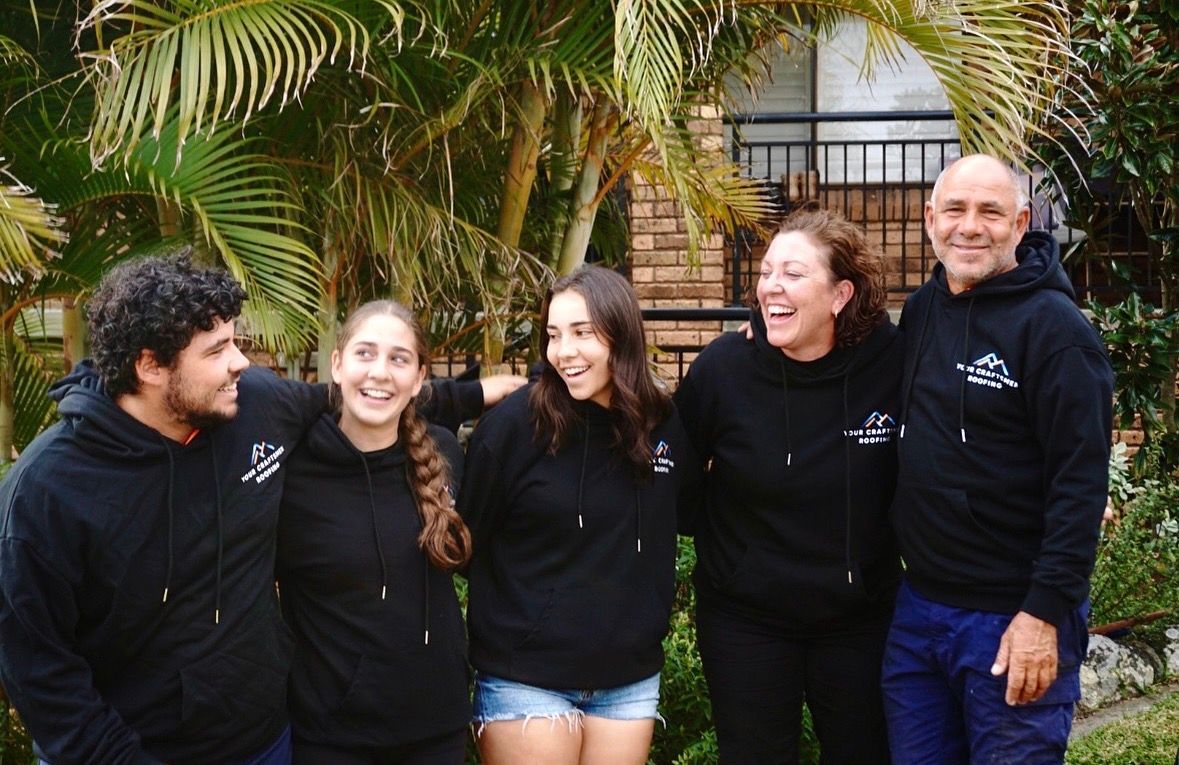 A group of five people stand outdoors, wearing matching black hoodies and smiling, set against a backdrop of green palms.