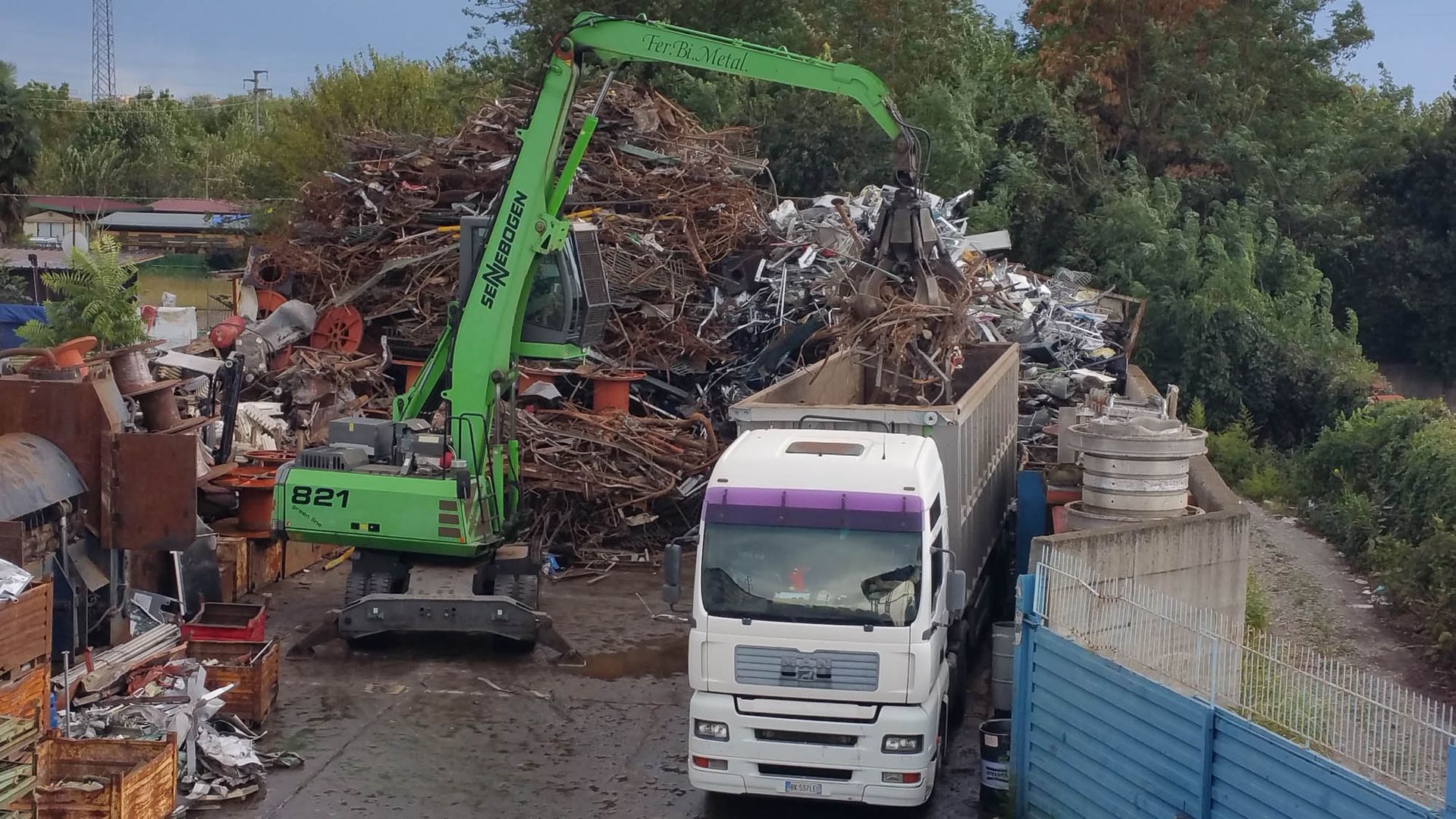 Un camion bianco viene caricato con rottami metallici da una gru verde.