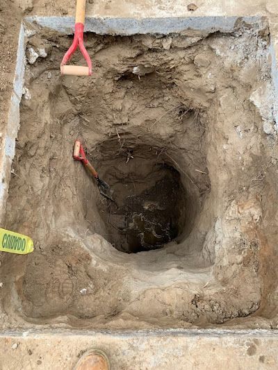 Top-down view of a square excavation pit with a shovel and red digging tool on the edge.