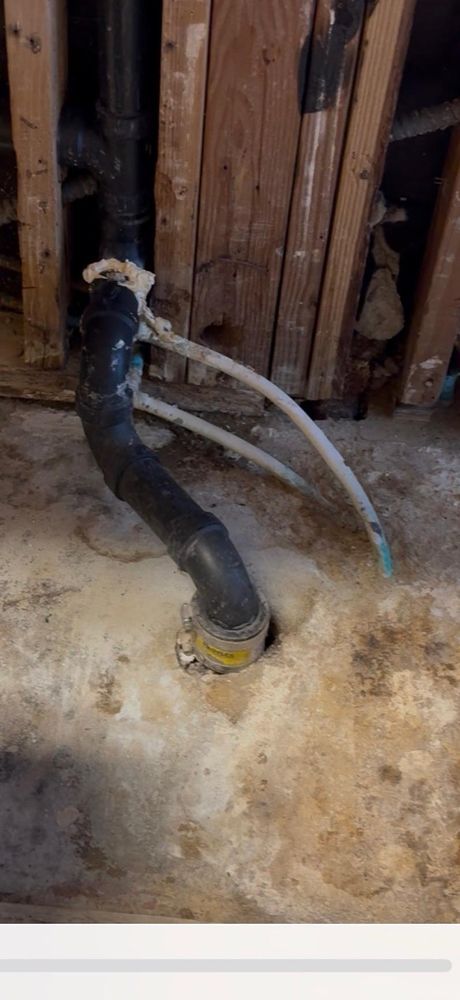 Black insulated pipe and two gray hoses rising from a floor opening in a basement wall.