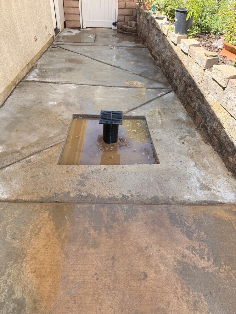 Narrow concrete walkway with a central drain pipe and water stains, bordered by a stucco wall and stone edging.