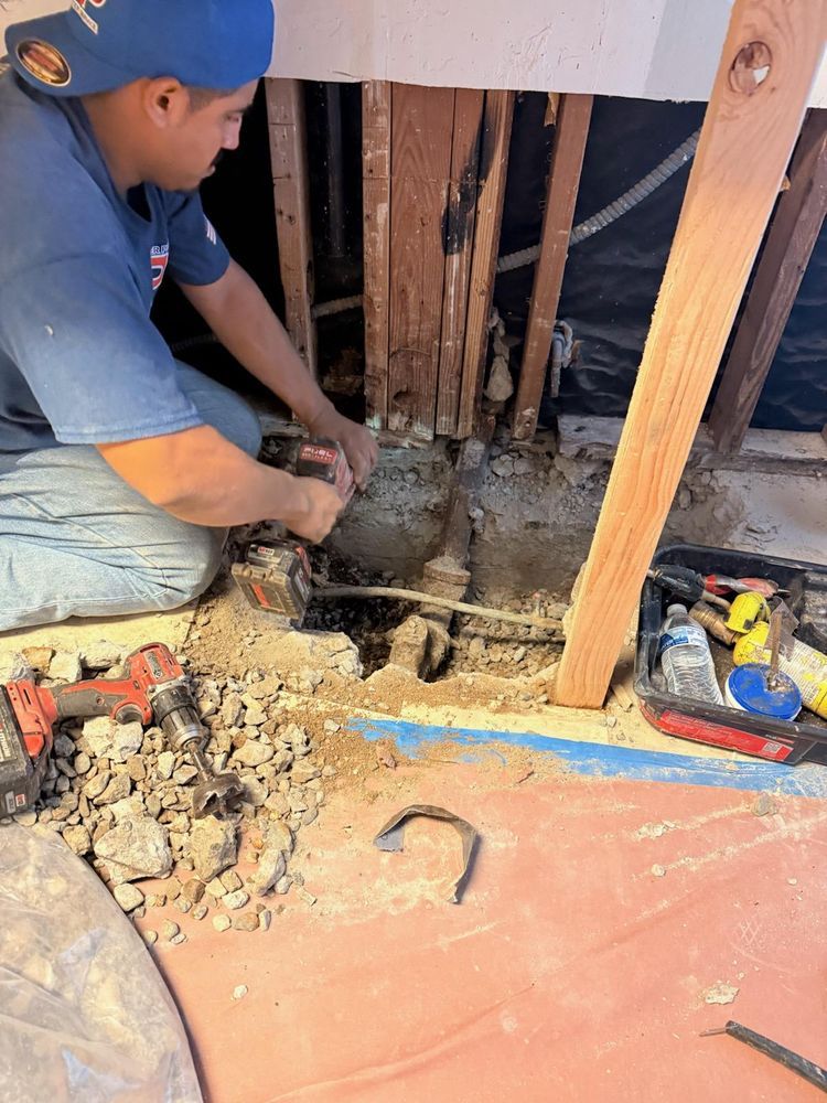 Worker kneeling beside a wall opening, repairing exposed pipes and debris on a construction floor