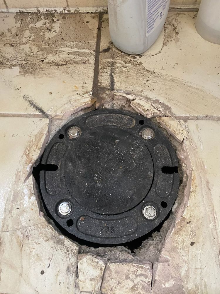 Black round sewer/drain access cover set into a stained concrete floor near a wall