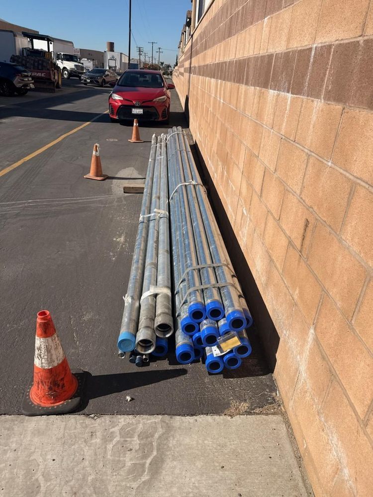 Metal pipes stacked beside a tan wall on a roadside, marked by orange traffic cones.