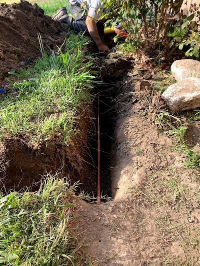 Person digging a narrow trench in a dirt path beside grass and rocks outdoors