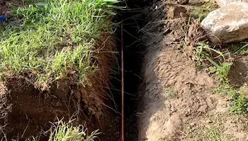 Narrow trench cut into soil beside grass and rocks, showing exposed earth along a yard edge