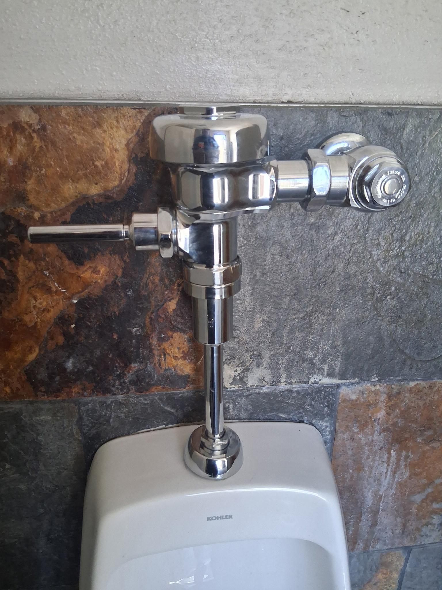 Chrome urinal flush valve mounted on a tiled restroom wall