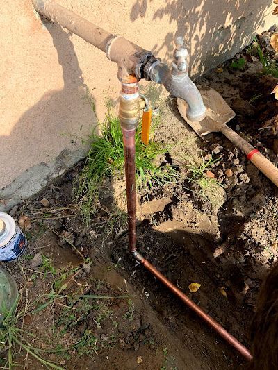 Outdoor plumbing repair with exposed pipes, a faucet, and a trench beside a wall.