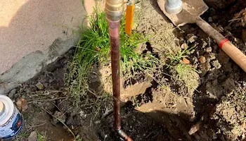 Rusty water pipe and muddy ground beside a house, with scattered tools and plants.
