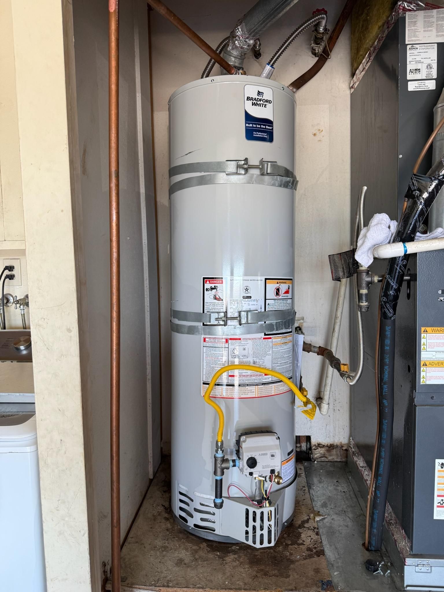 White water heater in a utility closet with exposed pipes and wiring