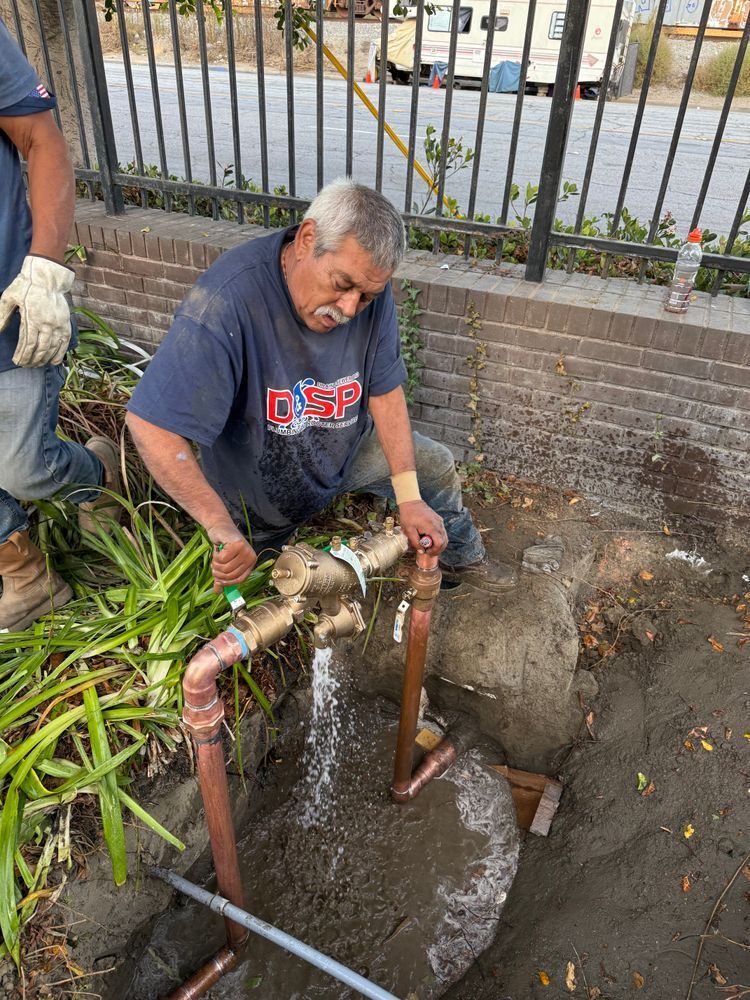 Worker repairing a broken water pipe beside a brick wall, using metal pipes and tools outdoors