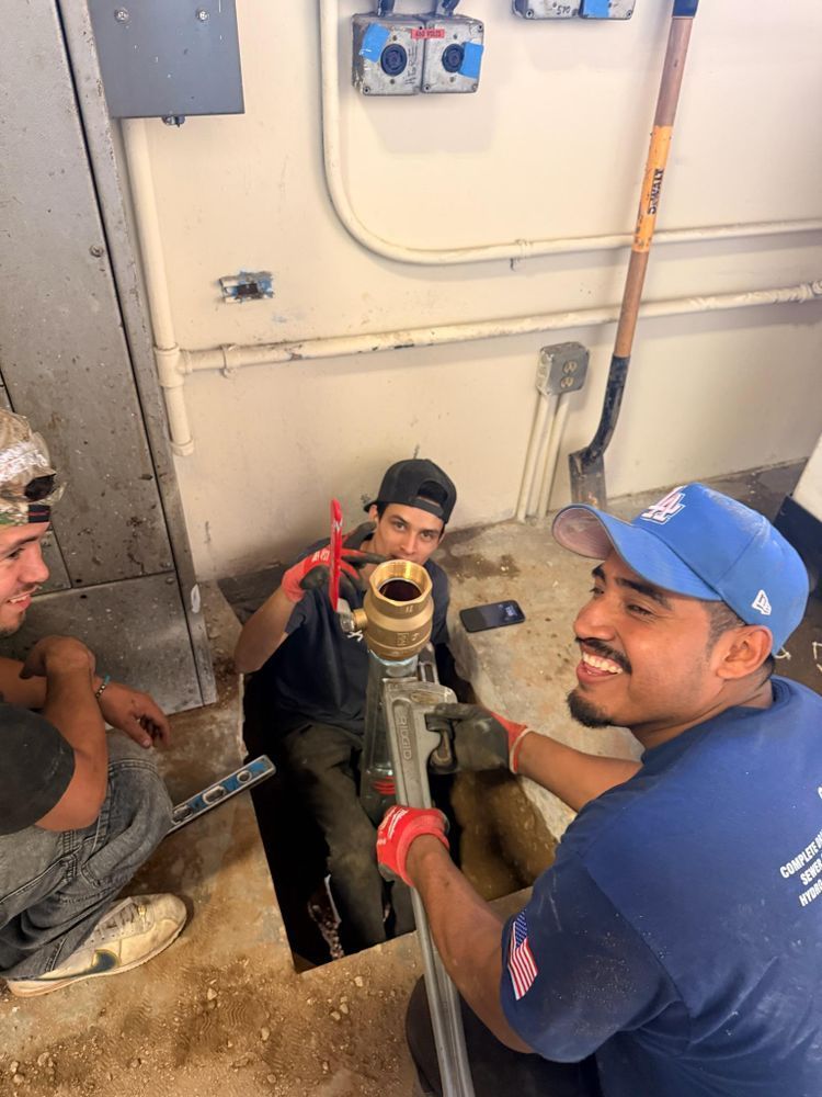 Workers using tools on a pipe in a utility room; one man in blue smiles while another uses red pliers.