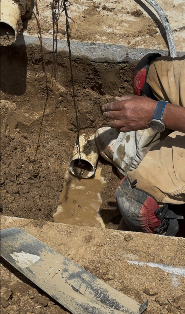 Worker crouches in a dirt trench, handling a pipe as muddy water flows out.