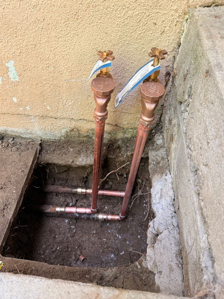 Two copper water taps in a concrete utility pit beside a wall, with a short pipe connecting them.