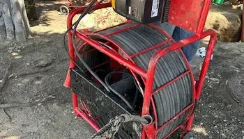 Red metal cable reel with coiled black hose on a dirt worksite