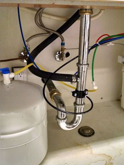 Under-sink plumbing with pipes, valves, and hoses connected to a white container next to a drain opening