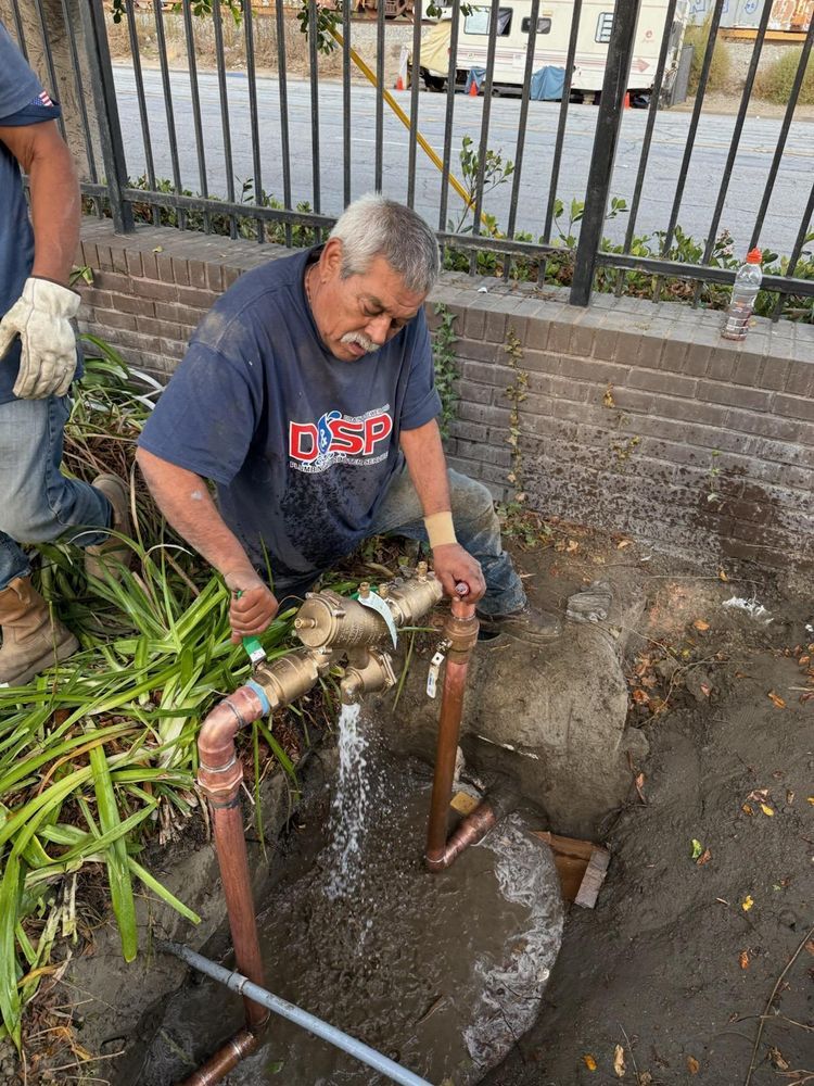 Worker repairing a water pipe in a muddy roadside trench near a fence