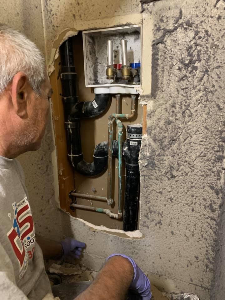 Technician inspecting exposed plumbing and electrical wiring inside a wall opening in a damaged room