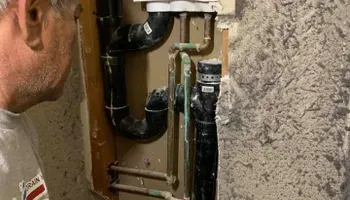 Plumber inspecting black pipes and a water meter in a basement utility area.