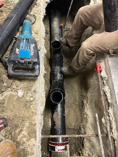 Plumber installing black PVC pipe in a trench beside a blue tool, with gloved hands visible