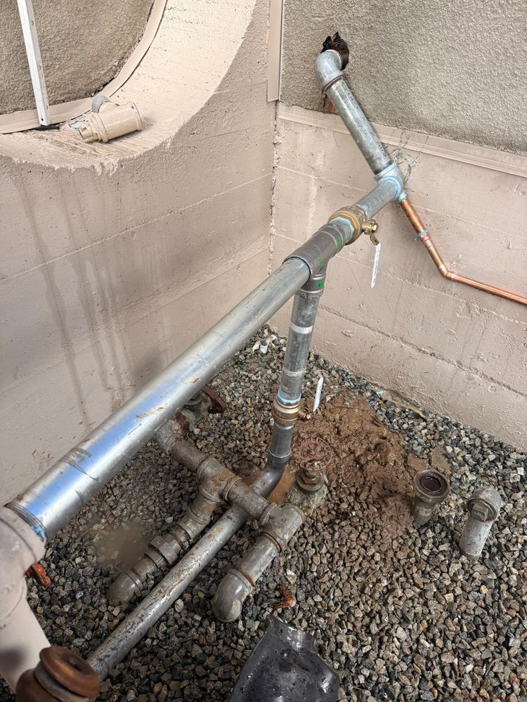Corroded outdoor plumbing pipes and valves beside a concrete wall and circular opening on gravel.
