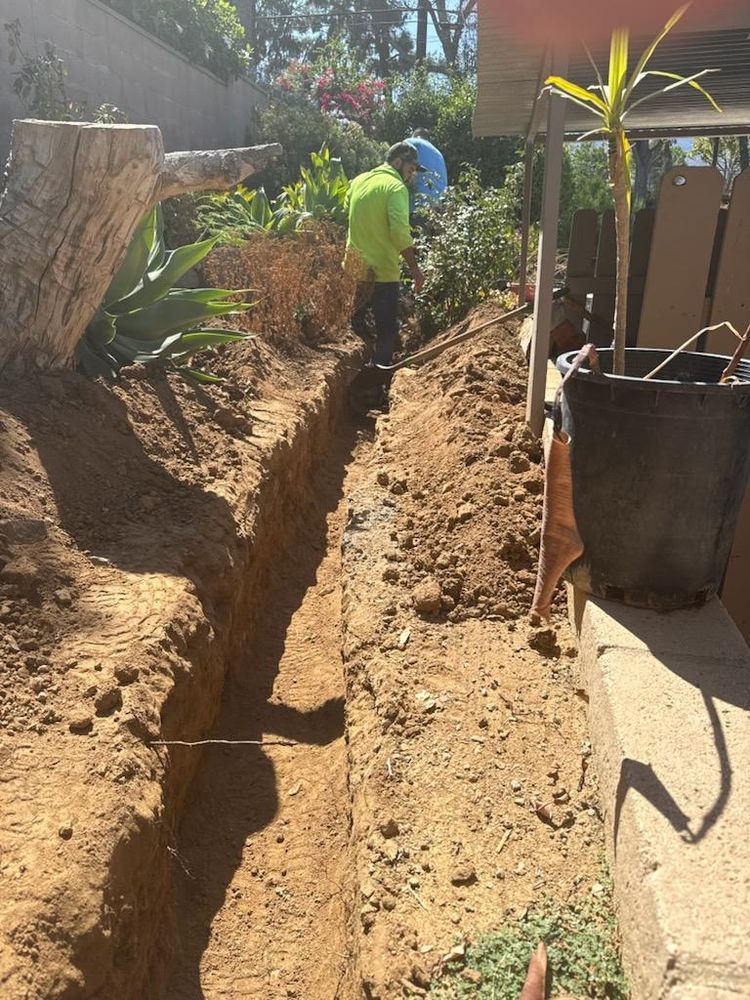 Workers digging a long trench in a dirt yard beside a house, with tools and potted plants nearby