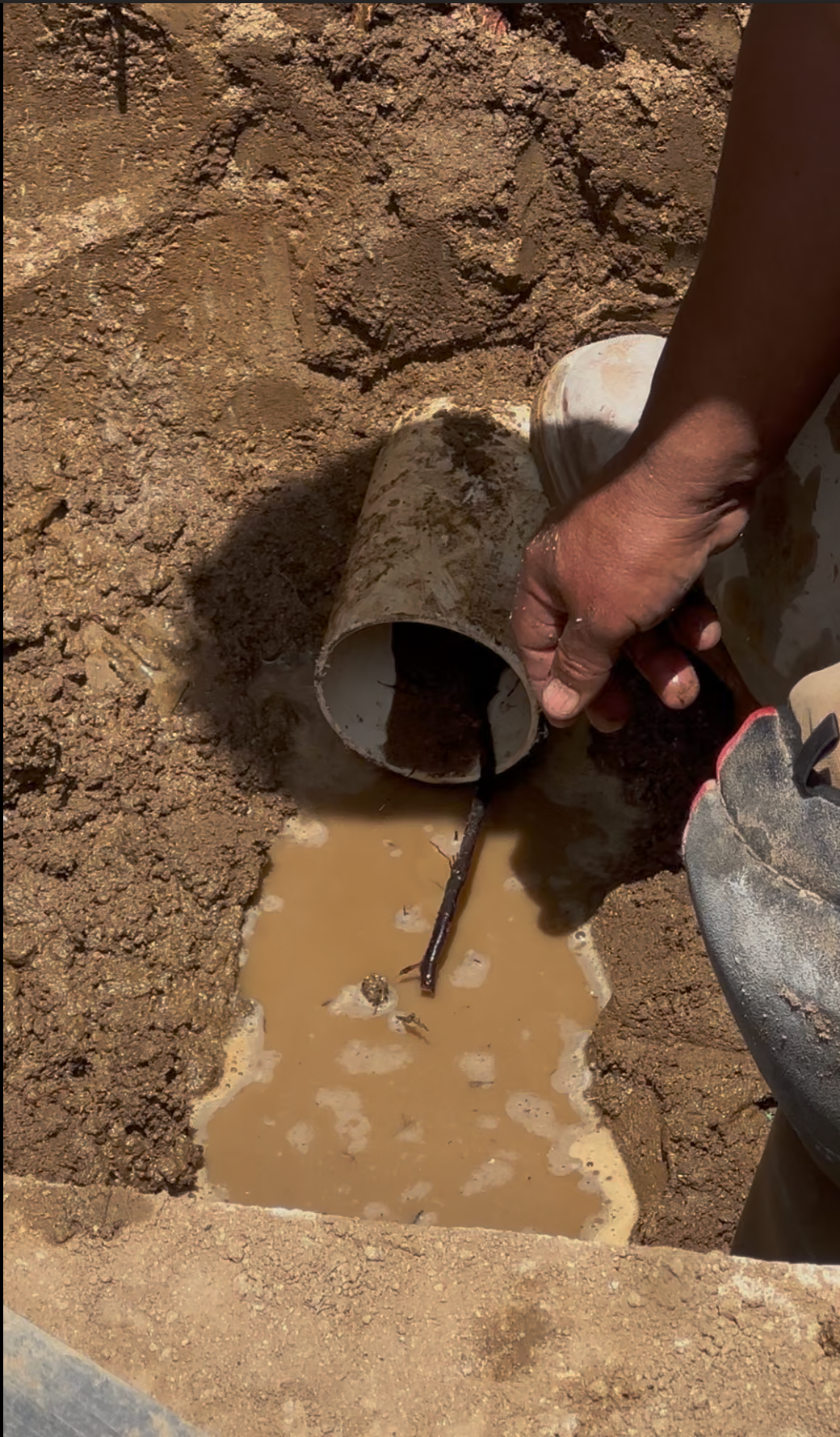 Hand pouring muddy water from a pipe into a small trench in a dirt excavation site