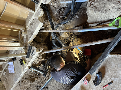 Worker installing plumbing under a floor, surrounded by exposed beams, pipes, and tools.