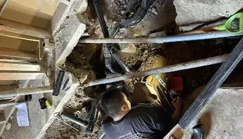 Worker crouching in a cramped basement, inspecting pipes and debris with a flashlight.