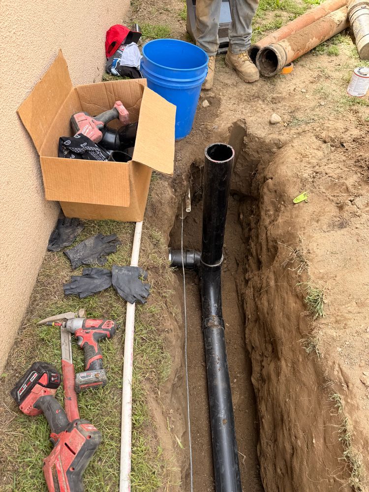 Utility trench with black pipe, blue bucket, tools, and a cardboard box at a construction site.