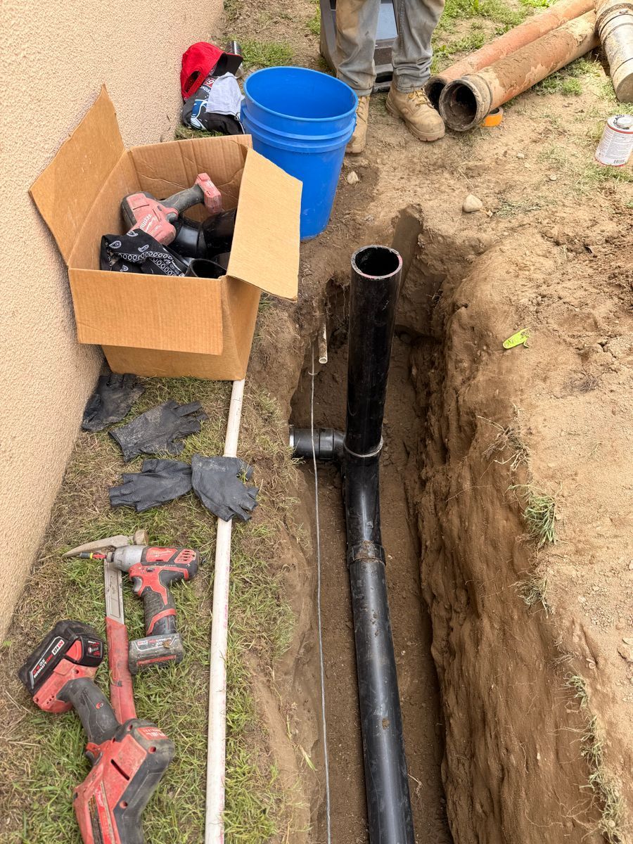 Open trench with black pipe, tools, gloves, and a blue bucket beside a cardboard box outdoors.