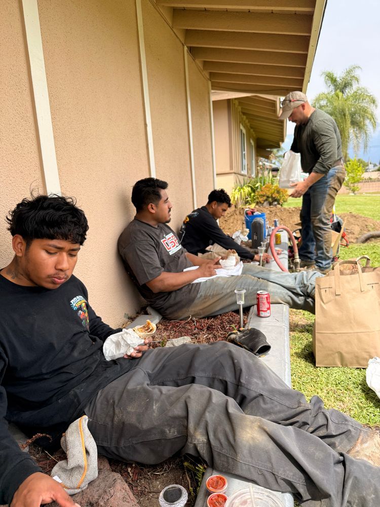 People sitting on a porch eating lunch, with one person standing nearby beside bags and drinks.