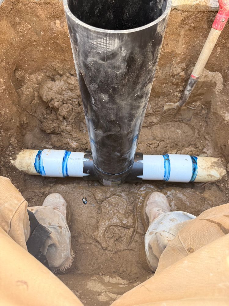 Worker inspecting a black pipe connected to a blue PVC fitting in a trench