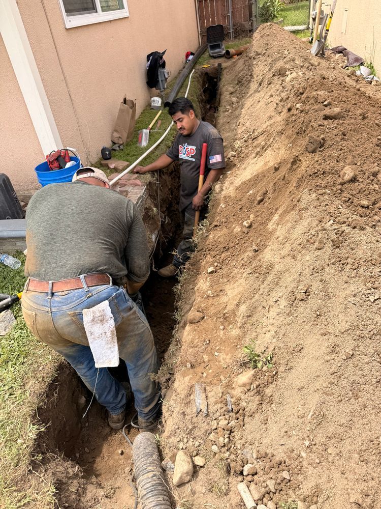 Two workers digging a narrow trench beside a house, with exposed dirt and utility pipes.