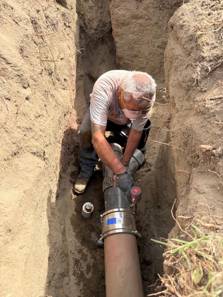Worker repairs a large underground pipe in a narrow dirt trench