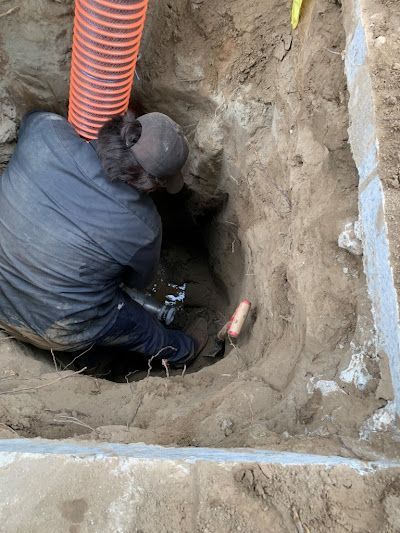 Worker in a trench using tools beside an orange corrugated pipe.