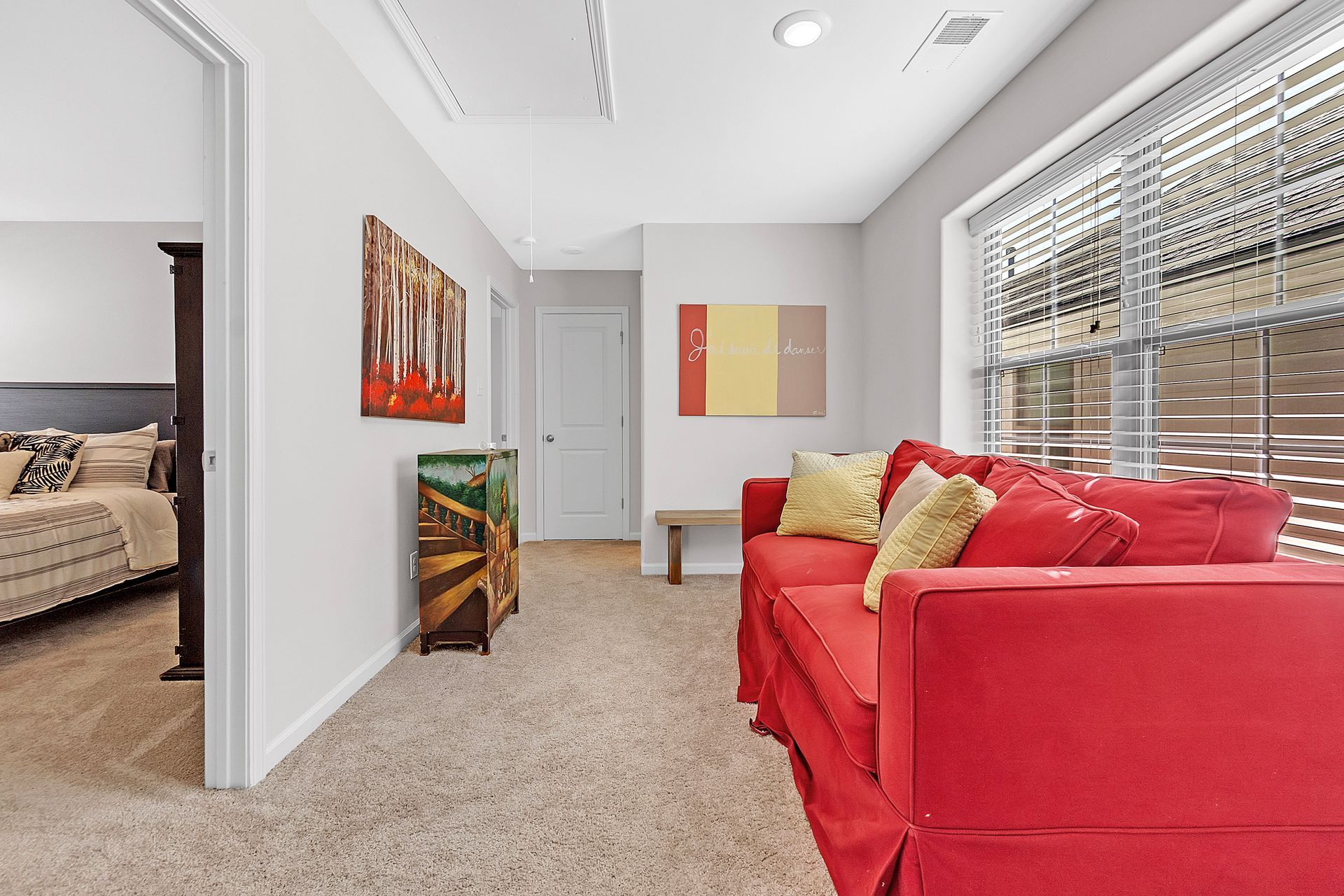Cozy living space with red sofa, art, window with blinds, and doorway to bedroom.