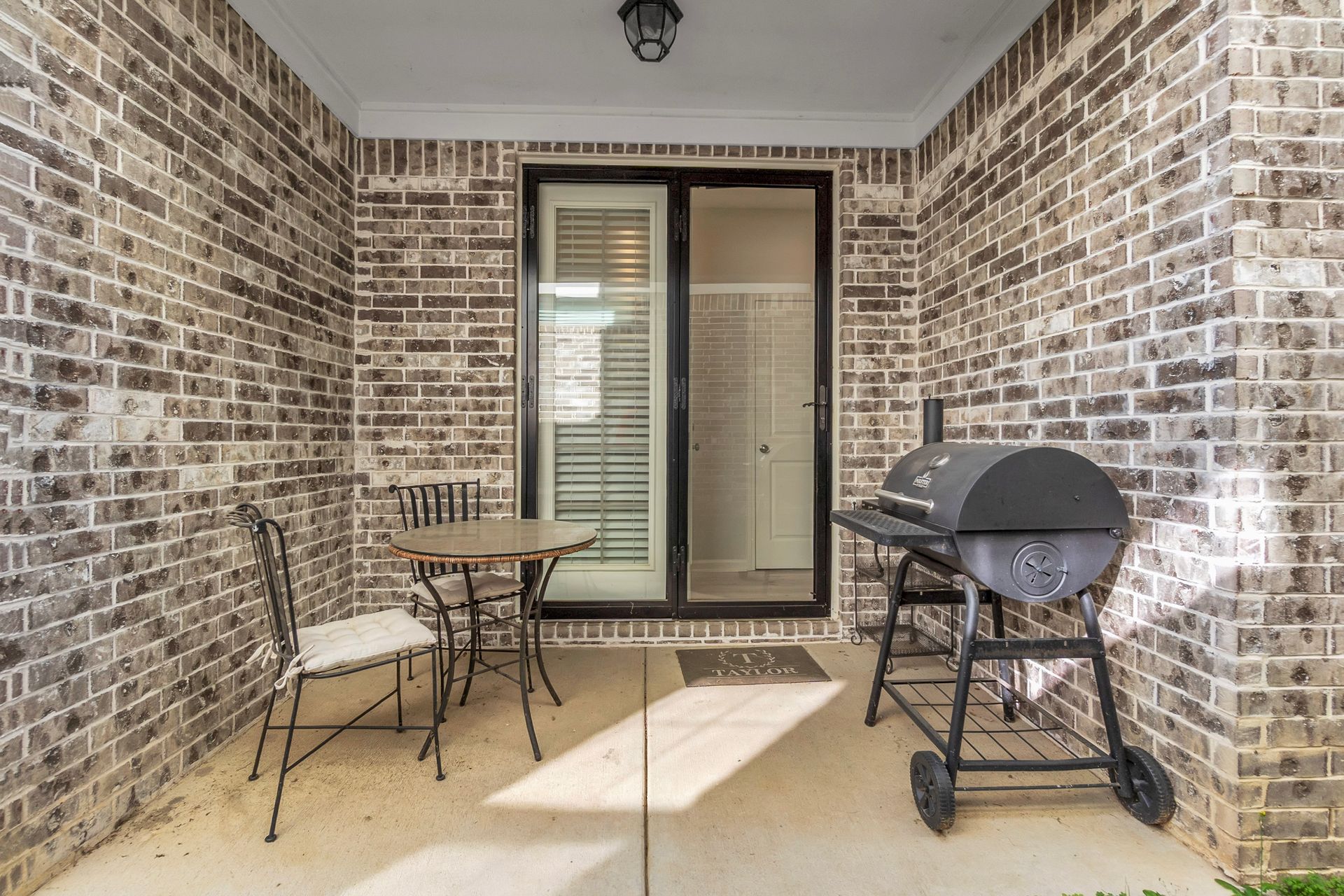 Covered patio with brick walls, small table and chairs, sliding glass door, and charcoal grill.