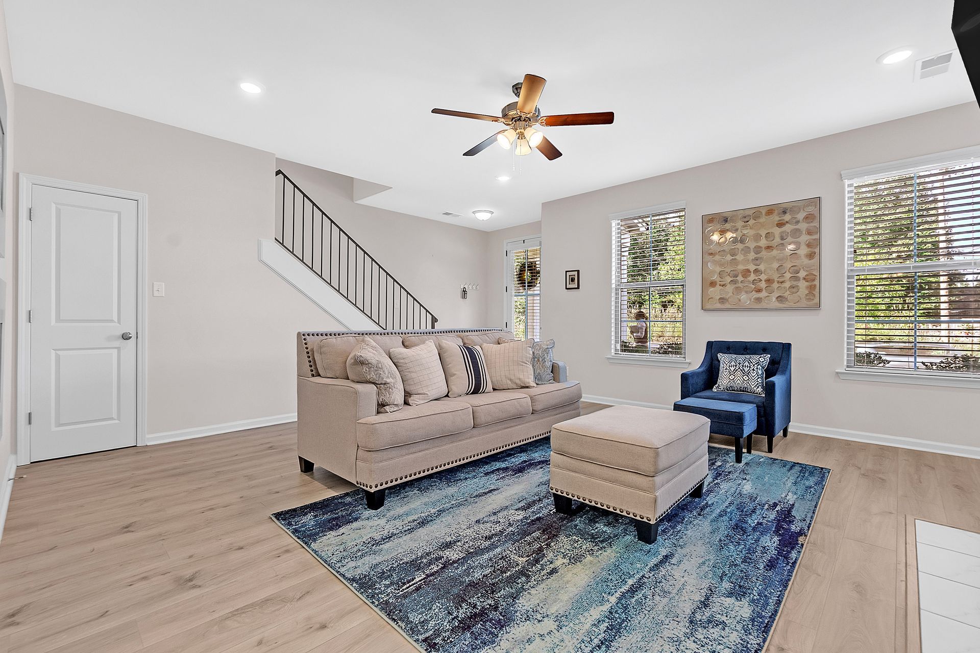 Living room with beige sofa, blue rug, and ottoman. Includes windows, staircase, and ceiling fan.