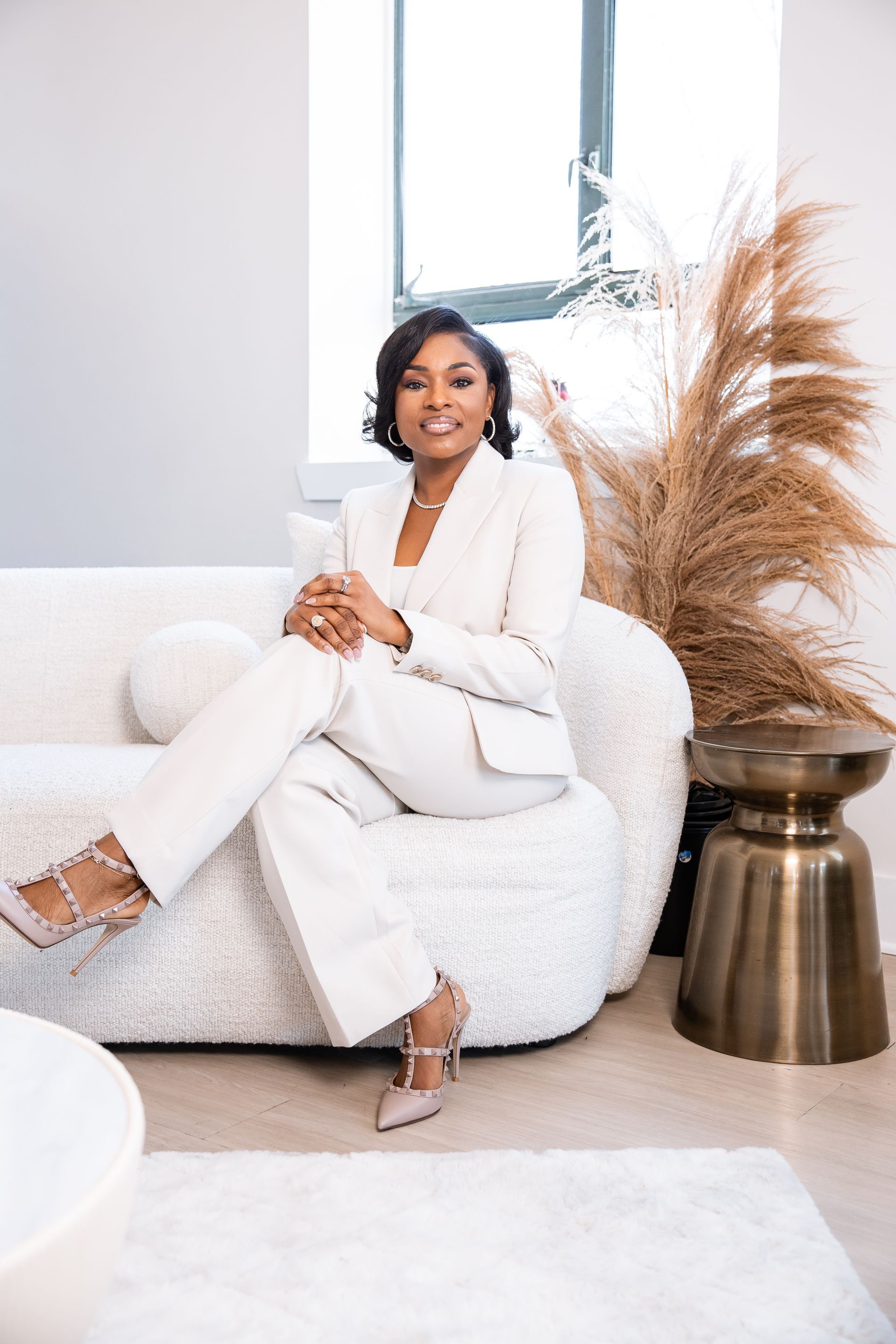 Woman wearing a beige pants suit, smiles, sitting on couch, hands folded over knee..