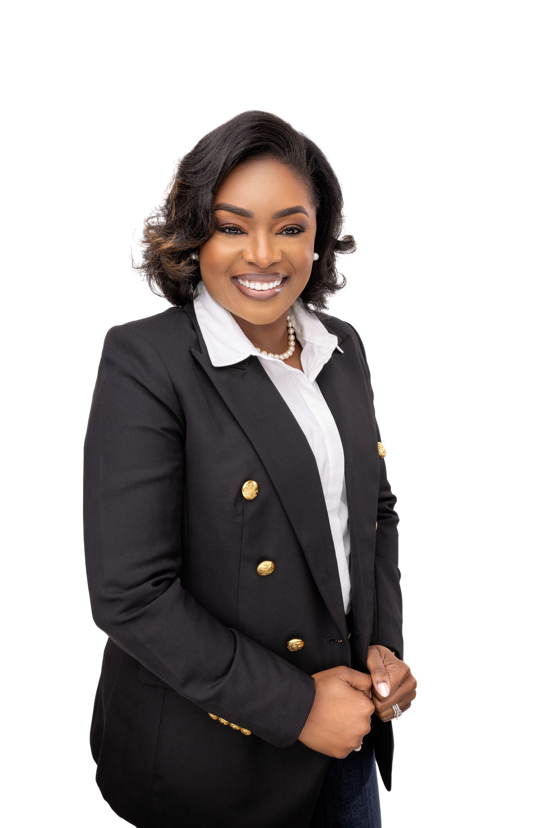 Woman in black blazer smiles, stands, hands clasped. White shirt and gold buttons visible.