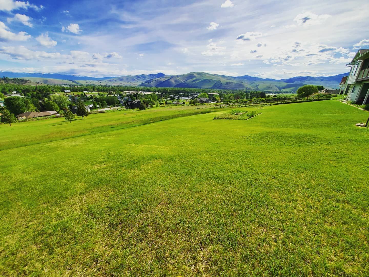 Green grassy field slopes toward a small town and mountains under a blue sky.