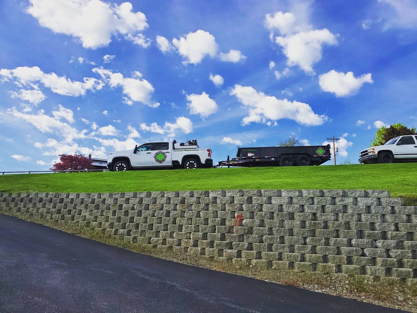 White trucks with trailer on grassy hill, gray retaining wall, blue sky with clouds.